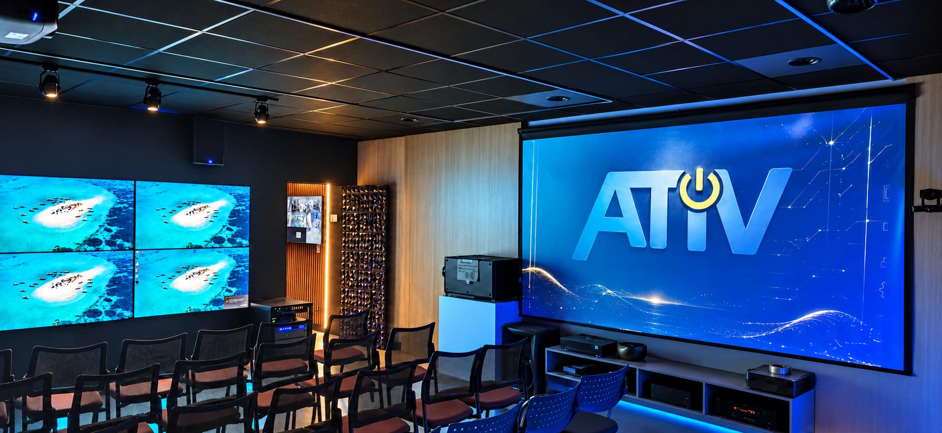 Sala de treinamento do AtivLabs com fileiras de cadeiras, videowall 2x2 à esquerda, tela de projeção exibindo a marca ATIV, projetor no teto e câmera PTZ
